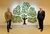 Commissioned by Age UK Two people dressed in suits standing either side a glass artwork mounted on a white wall. The artwork is a 2D glass tree with a brown trunk and disjointed limbs in between large clear pieces of glass that hold individual leaves in different shades of green.