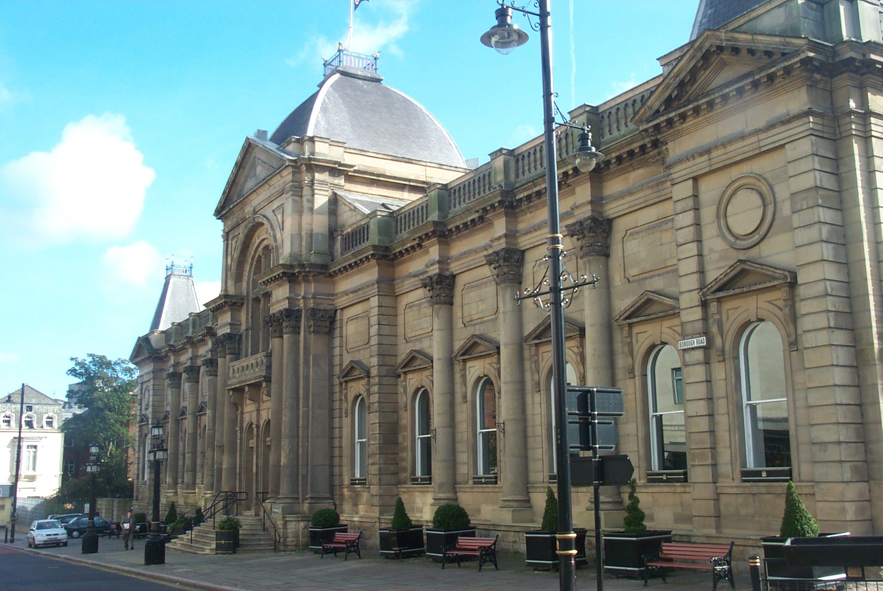 A large, historical, stone building with slim arched windows and a curved stone staircase leading up to two dark wooden doors at the centre. A domed grey roof at the top of the building has a white pole and a United Kingdom flag attached to it. There are wooden benches and black and gold plant pots at regular increments along the front of the building.