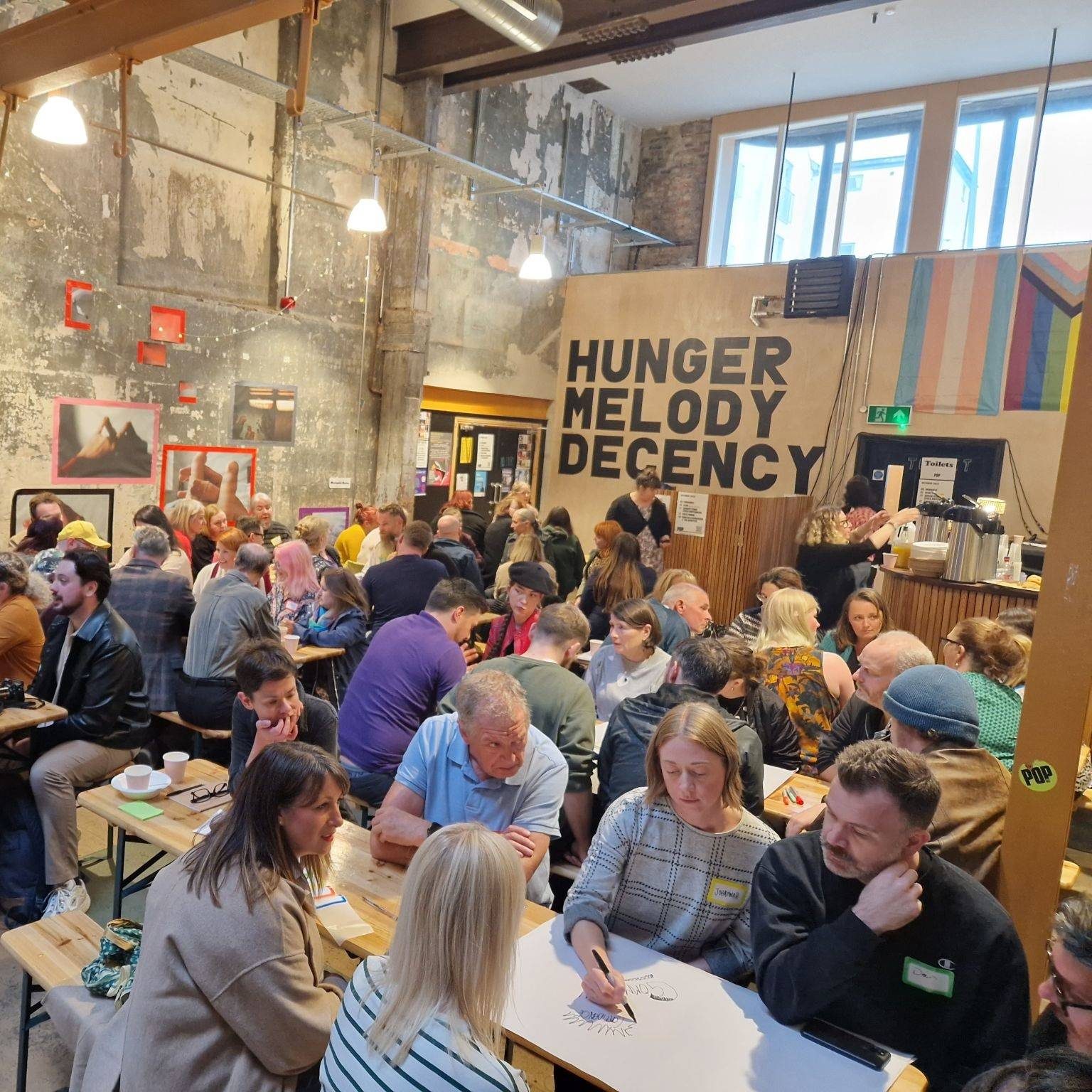 Groups of people sit at long wooden tables talking amongst one another with coffees, large sheets of paper and pens. The room has bare concrete walls and a yellow beam through the middle. At the back is a plywood wall that has large black text on it reading 'Hunger Melody Decency'.