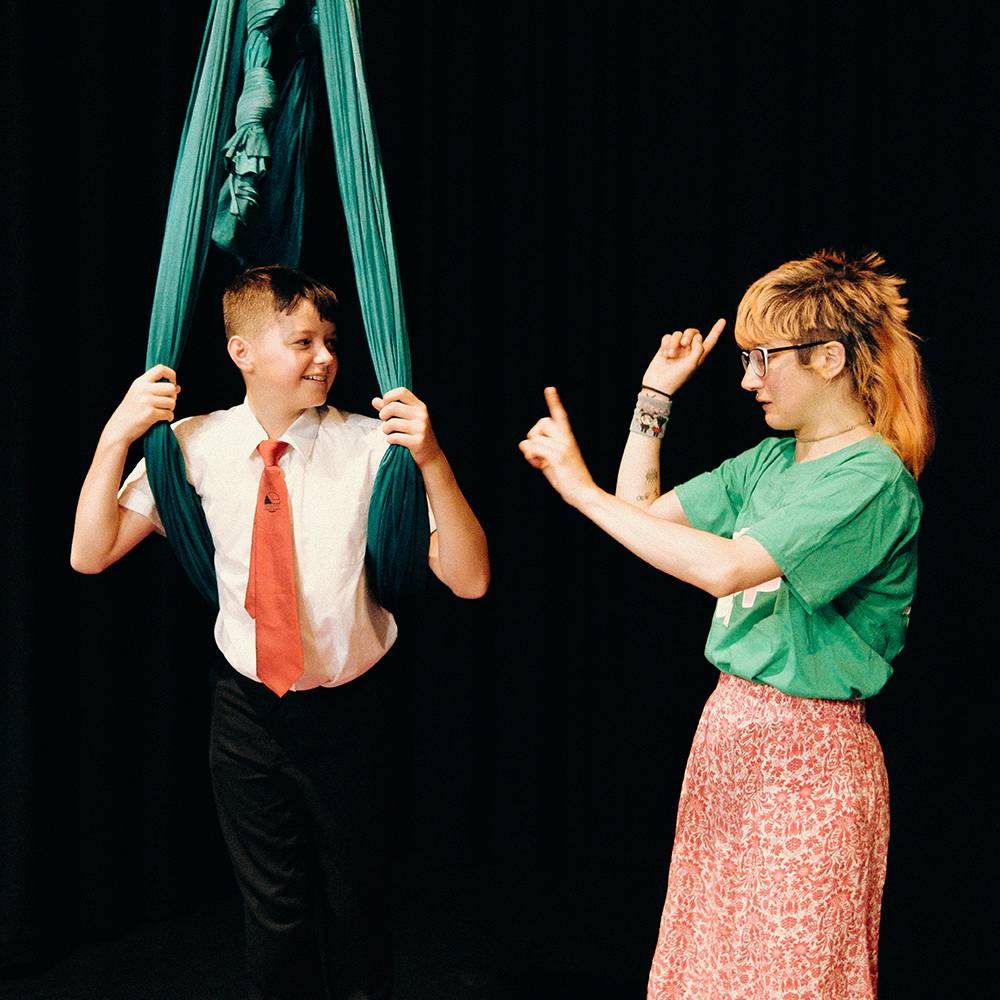 A young person wearing a white shirt and red tie, stands in a dark room with a green acrobat ribbon wrapped around his back and arms. Another person stands to their right talking and gesturing with their hands.