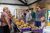 Craft fair at Arts Centre Washington A long table stacked with plates of pastries and other baked goods in a stone-walled room. Two people stand behind the table wearing aprons and plastic gloves taking to a group of people smiling and looking at the items on the table.