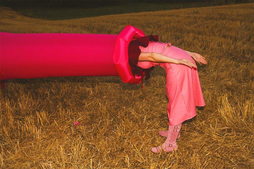 A barrel of a cannon points from left to right. The whole cannon has been covered in a shiny pink cloth. A female, also dressed completely in pink, bends at a right angle and sticks her head into the cannon. The cannon and the female are standing on very dry grass.