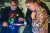 Autonomy Special Interests Club - Sunderland Culture Two individuals are engaged in an interactive activity using a tablet mounted on a stand. One person, wearing a floral-patterned shirt and glasses, leans in with an expression of curiosity and engagement. The other, wearing a dark cardigan over a colorful dinosaur-print shirt, gestures toward the tablet screen, seemingly explaining something. The background is softly blurred, with other people and objects visible in the space, suggesting a hands-on or educational environment.