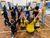 Culture Start School Activities | Sunderland Culture A group of children and an adult stand or kneel in a school hall, facing away from the camera with their arms raised. They wear handmade, colorful dragon wings attached to their backs, featuring different patterns and drawings. The children wear navy and yellow uniforms, while the adult wears a mustard-yellow top and jeans. The hall has a wooden floor, blue chairs stacked against the wall, and motivational words displayed on the walls.
