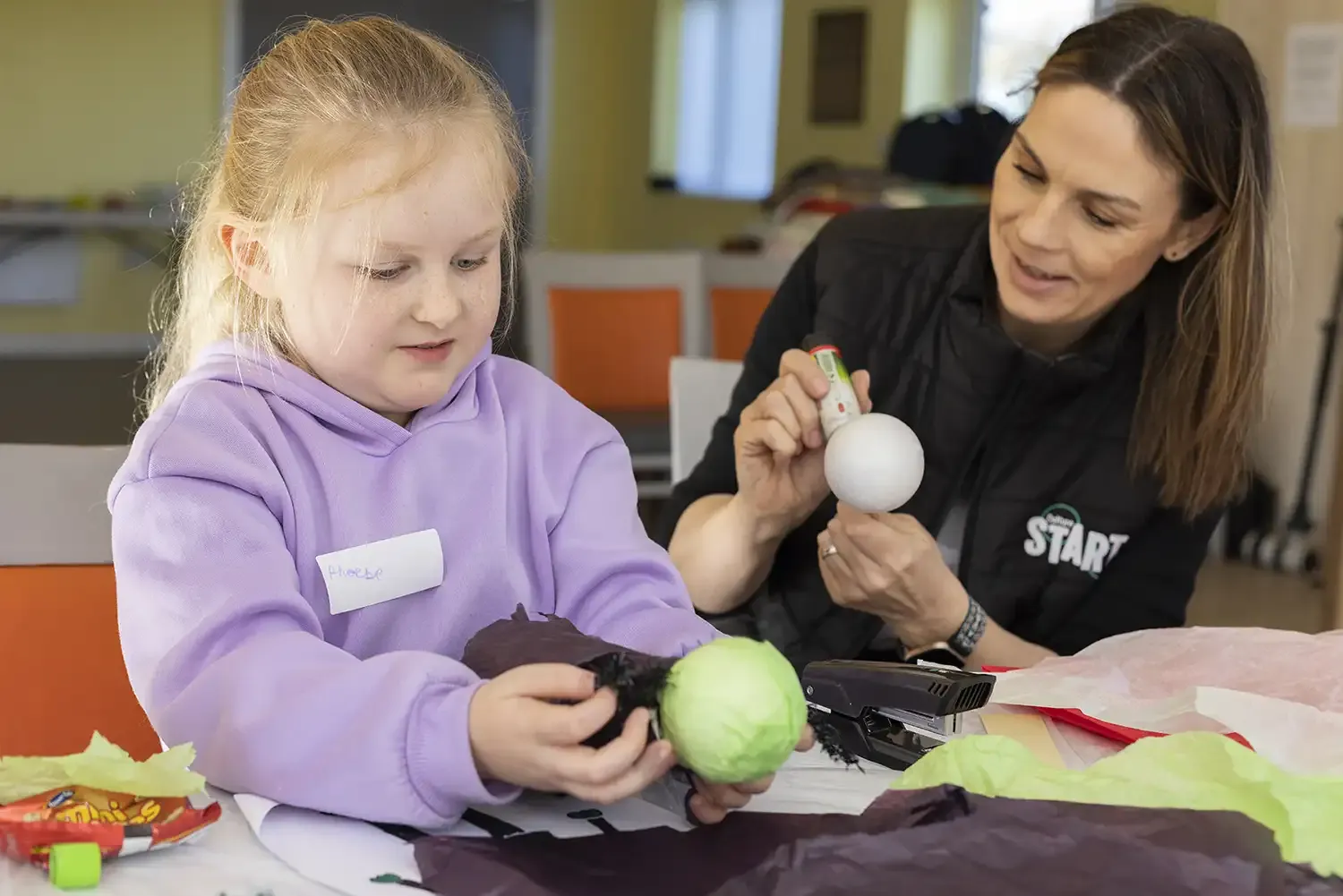 Two people working on a craft project at a table. On the left is a young person with light hair wearing a purple hoodie with a name tag. This person is holding a green ball and some dark fabric. On the right is an adult with long hair wearing a black top with 