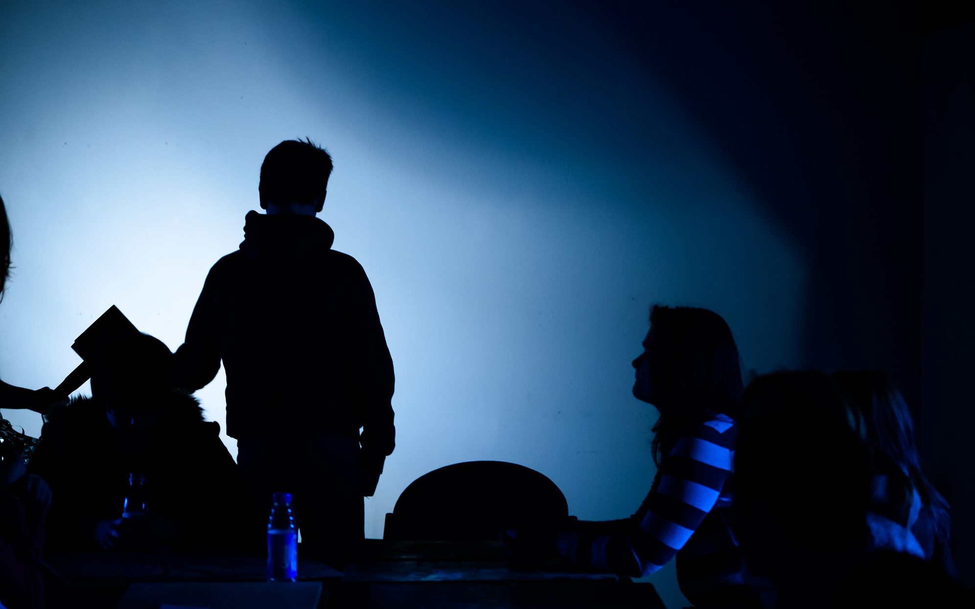 A dimly lit scene with deep blue lighting. Several people are silhouetted against a blank wall, with one standing and others sitting at a table. The mood is tense or mysterious, suggesting a theatrical performance or dramatic workshop