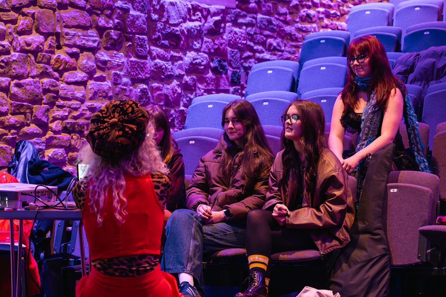 A small theatre or performance space with purple lighting. Five young people are sitting in blue theatre seats. One young person with red hair and glasses stands on the right. Three others sit in the front row wearing dark jackets, and one has colourful socks. A person with curly hair in red clothing sits with their back to the camera on the left. The background shows a stone wall lit with purple light. Many empty blue seats are visible behind the group. The group appear to be having a conversation or engaged in a small seminar.