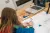 Animation workshop at Bright Lights Youth Arts Festival A child with long light brown hair, wearing a blue fury jacket, sits at a wooden table drawing on a large piece of paper. At the top of the frame you can see an open laptop and a short, black tripod.