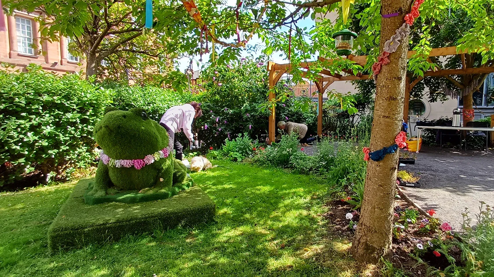 A sunny garden with bright green grass and flower beds. In the centre sits a large topiary sculpture shaped like a frog with a pink flower garland around its neck. A person in a white shirt tends to plants in the background. Colourful ribbons and decorations hang from tree branches overhead. Traditional brick buildings with white window frames can be seen behind the garden. Wooden garden structures and planters are scattered throughout the space, creating a cheerful community garden atmosphere.
