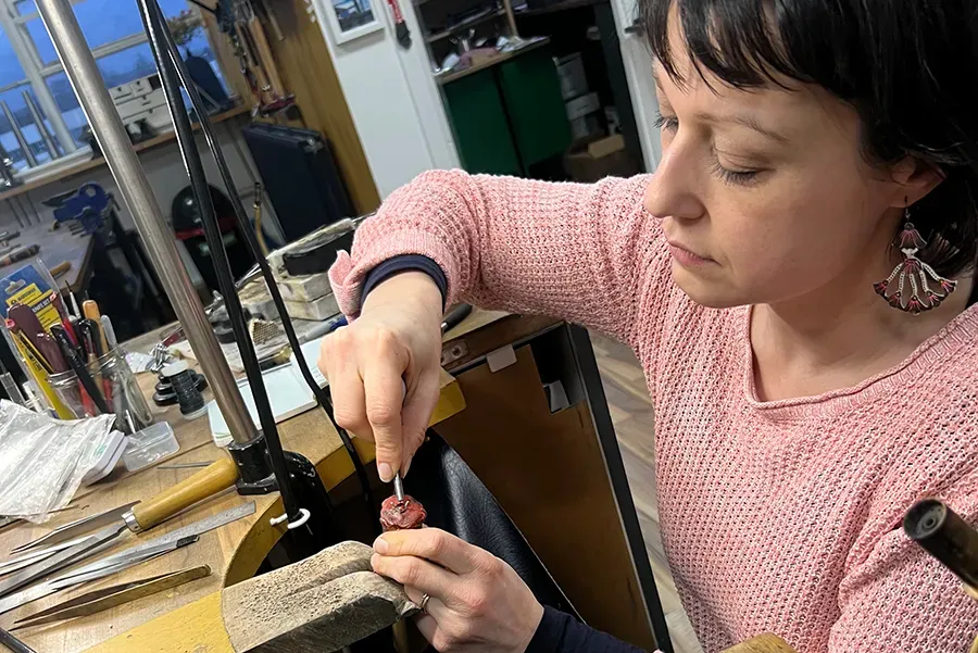 A person wearing a pink jumper with short brown hair sits at a wooden work bench, working on a small piece of metal. The workbench has a variety of metal tools. There is another workbench and larger tools in the background.