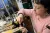 Artist Joanna Manousis, Unlock Grant Recipient A person wearing a pink jumper with short brown hair sits at a wooden work bench, working on a small piece of metal. The workbench has a variety of metal tools. There is another workbench and larger tools in the background.