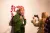 Autonomy Gallery Visit A person with blond hair, wearing a green jacket and lanyard, poses enthusiastically with two thumbs up in front of a wall with red symbols. Another person with long blond hair and a fluffy coat takes their photo with a smartphone.