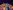 The Odyssey - Episode 4: The Island of the Sun at The Fire Station A person with curly hair and glasses stands centre stage with both arms raised high in triumph. They wear a purple top and green trousers. Around them, other performers in colourful clothes cheer and clap. Purple stage lights shine down from above, and a large yellow circle glows behind them on the stage backdrop.