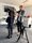 Film making at Back on the Map Two people work with camera equipment in an office room. A person with long brown plaited hair wears a dark blue hoodie and grey trousers, holding a large black camera. Behind them, someone in a grey shirt helps adjust the camera setup. The room has grey carpet, white walls, and a large TV screen on the wall. There are camera tripods, white desks, and storage cupboards around the room. A microphone on a stand sits nearby.