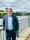 Nick Malyan, Chief Executive, Sunderland Culture A person with short grey hair and glasses stands smiling on a riverside path. They wear a blue jacket over a light blue shirt with dark trousers. Behind them is a river with a green metal bridge crossing it. Grey and white clouds fill the sky above green trees and tall buildings in the distance.