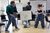 Our Freedom Young People's Workshop Three young people perform a drama activity. Two stand on either side holding up a large white paper with drawings of a face and book. A third person kneels behind the paper, creating a puppet-like scene. Black chairs are arranged around them in the bright classroom.