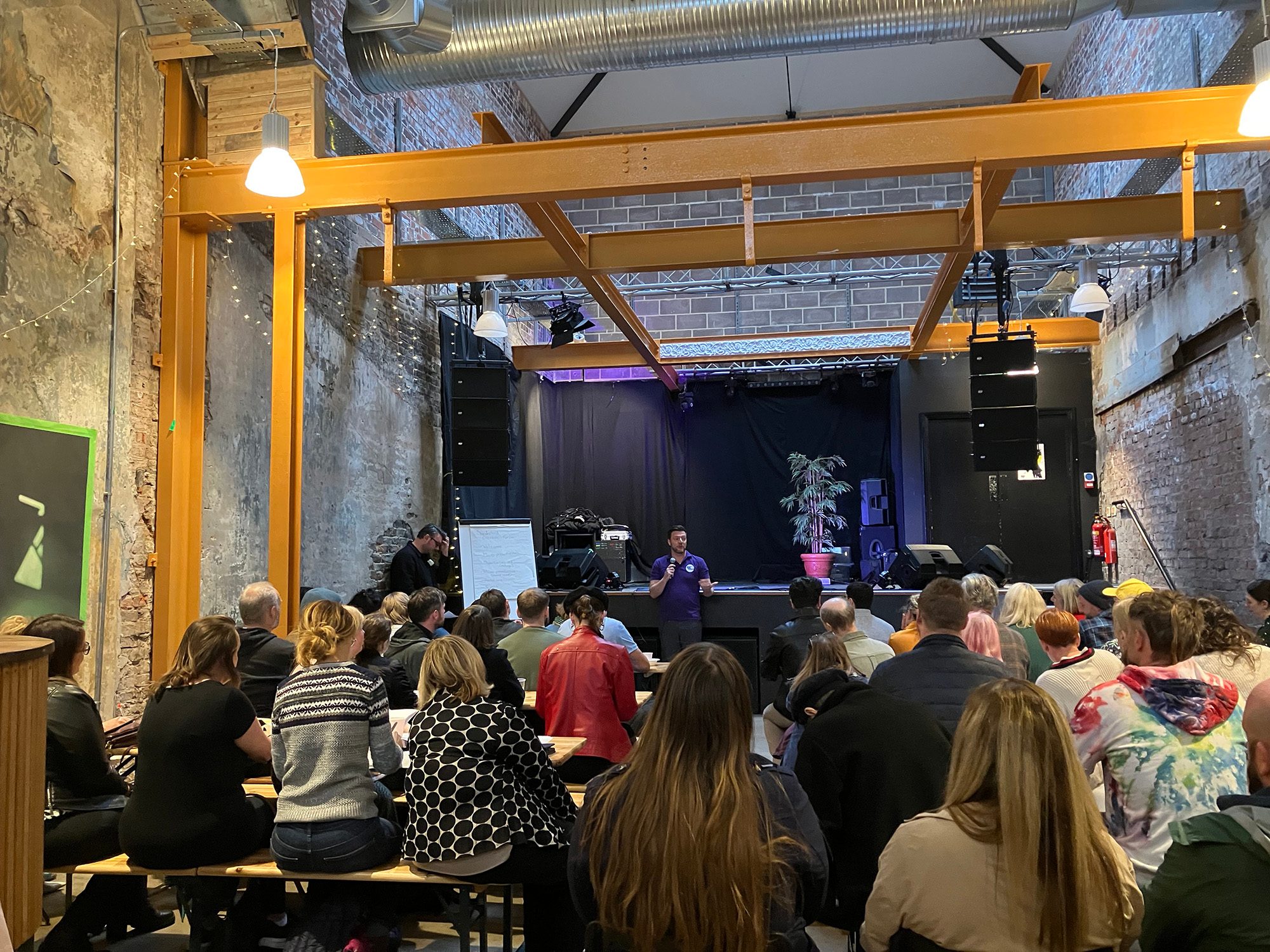 A wide shot of an indoor event space with a high ceiling. A group of people are sitting on benches and facing a small stage area. On the stage, a person is speaking into a microphone. The room has exposed brick walls and large yellow metal beams across the ceiling.