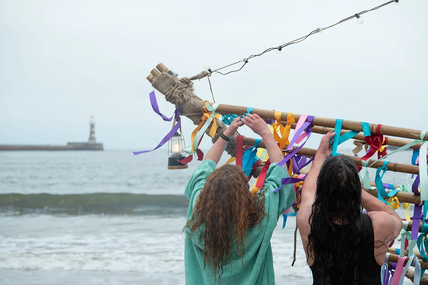 Two people stand on a beach with their backs to the camera. They tie many colourful ribbons to a long wooden pole. The ribbons are purple, blue, yellow, and red. A small glowing lantern and a string of lights hang from the pole. In the background, the sea has small white waves. Far away in the water, there is a tall white lighthouse on a long stone wall. The sky is grey and cloudy.