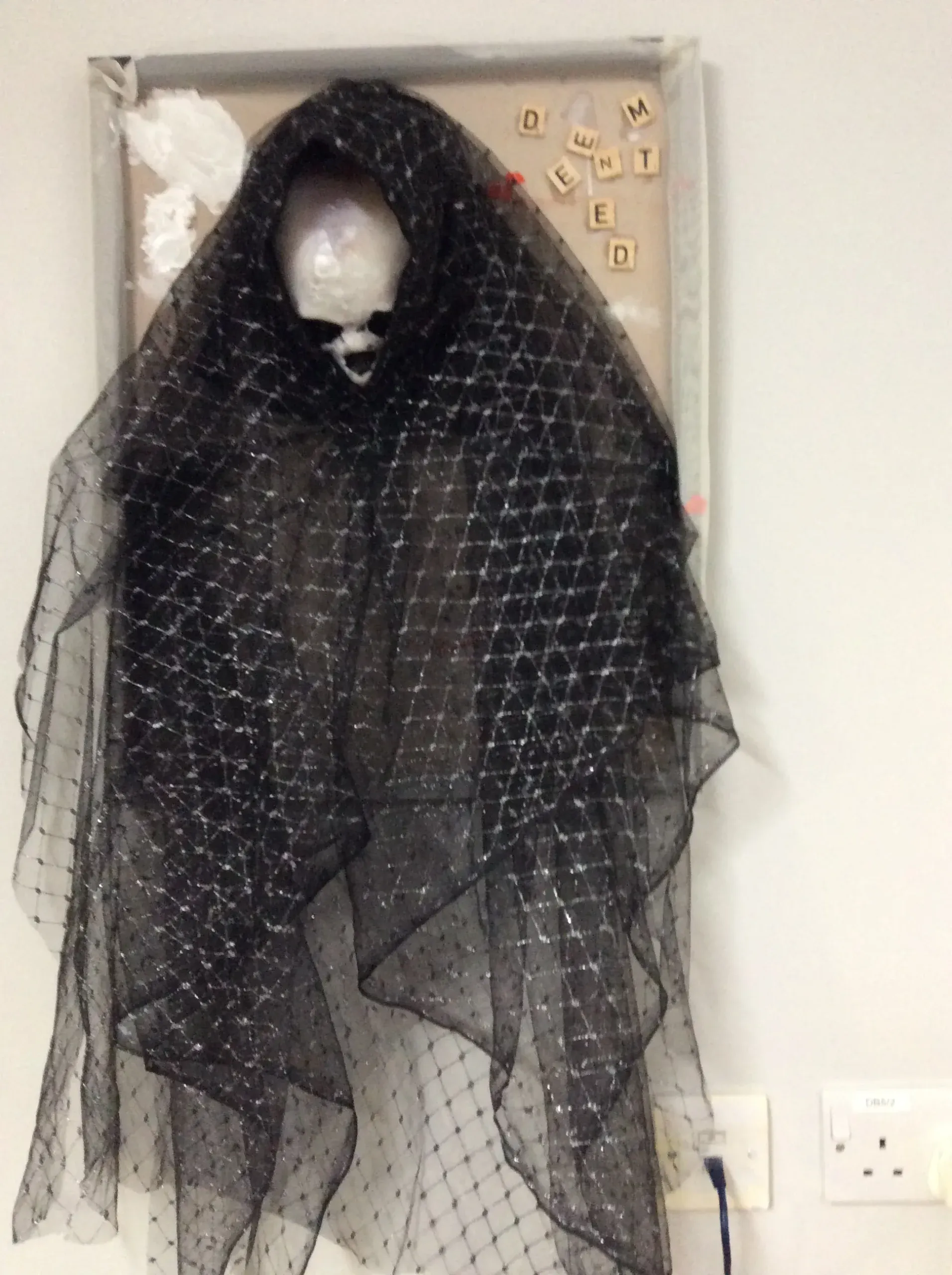 A white skull hangs on a piece of cork board with a grey frame. Black mesh cloth with a silver diamond pattern is draped around the skull. To the right of the board, small wooden tiles placed in a haphazard cluster appear to spell out 'Demented'.