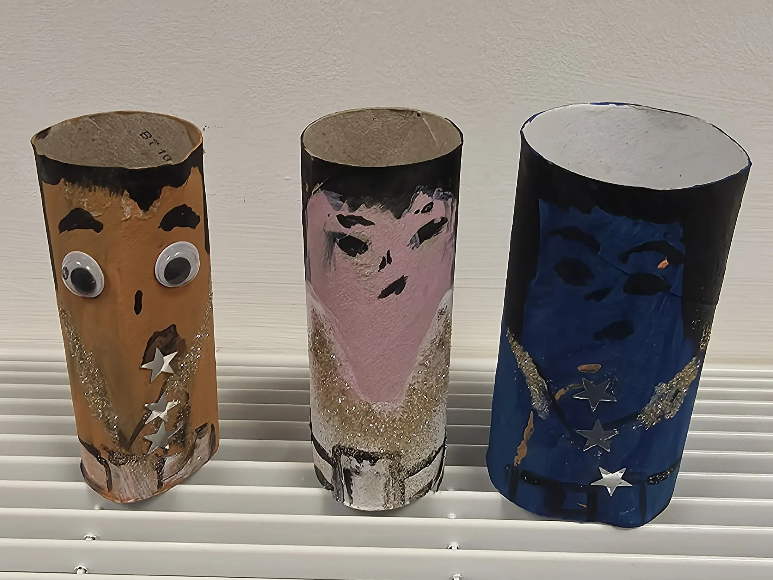 Three decorated cardboard tubes sit on a white ribbed surface. Each tube is painted to look like a person with a different outfit. The one on the right is painted brown with large googly eyes and has silver stars stuck in a line down the middle. The middle one, is painted pink with a simple face in black. The outfit is white with gold glitter in a 'V' shape. The one on the right is slightly larger than the others, painted blue with a simple face in black. This one has silver stars down the middle and a patch of silver glitter on either side.