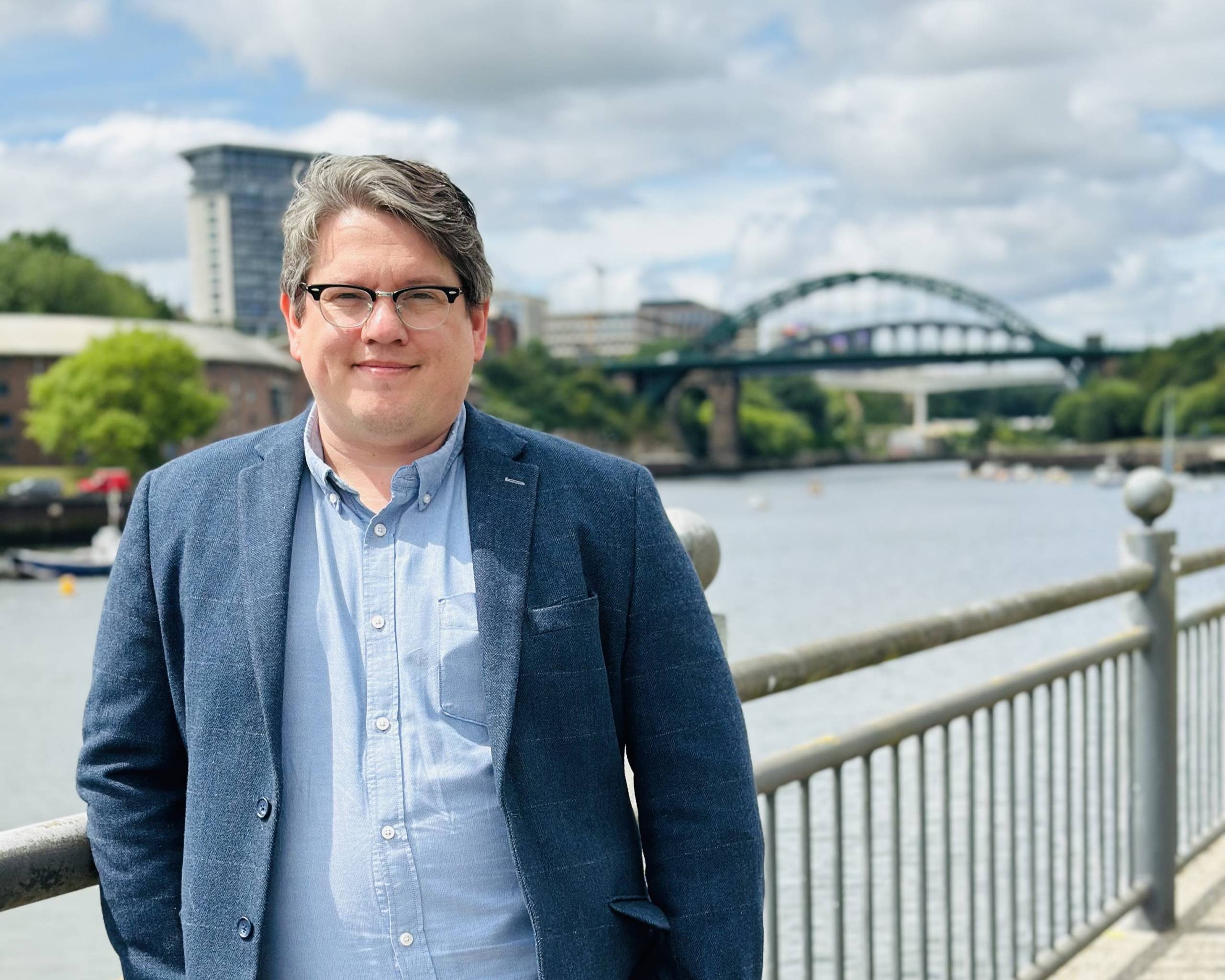 Man wearing glasses and a blue blazer standing by a riverside railing, with a large green arch bridge and city buildings in the background.