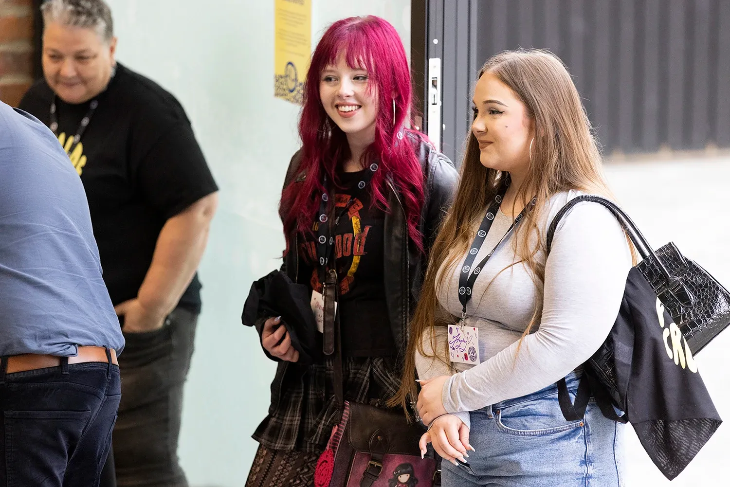 Two young people stand together, smiling and looking toward something out of frame. The person on the left has long, vibrant magenta hair, wears a black graphic t-shirt under a black leather-style jacket, and a plaid skirt. The person on the right has long brown hair and wears a light grey long-sleeved top and blue jeans, carrying a black tote bag. Both are wearing lanyards with ID badges. In the blurred background, another person in a black t-shirt and a person in a blue shirt are partially visible.