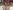A colourful model made from a K'NEX kit resembling a windmill sat on a white table. Behind the model, on the table is a grey tray surrounded by colourful plastic blocks and in the background is a pink archway set in a wall of glass looking out onto a darkened corridor.